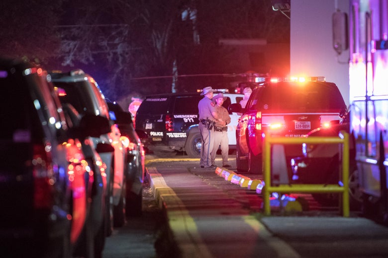 A number of police vehicles with lights can be seen. A small group of officers are gathered for a conversation in the distance.